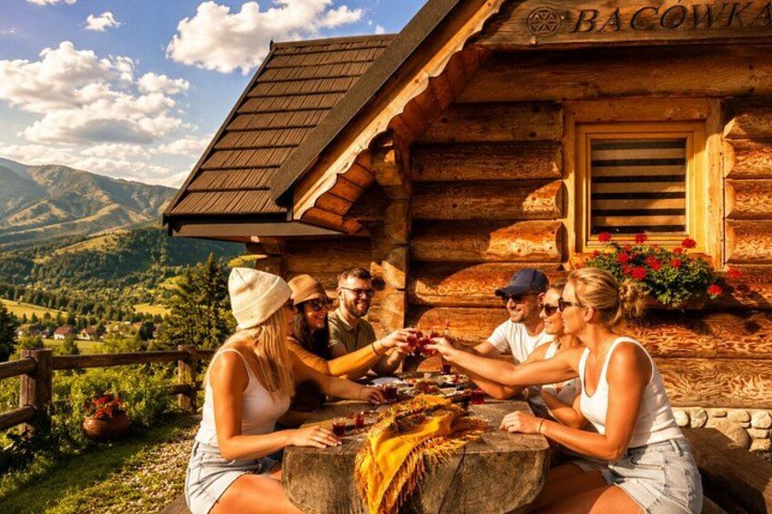 A wooden cabin with a sign reading 'BACOWKA' and a group of people sitting at a table outside. 5