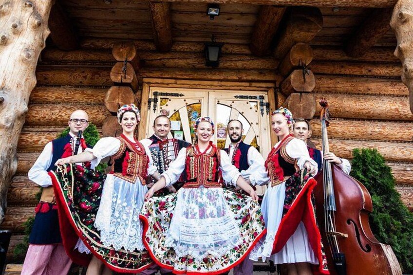 A group of people in traditional Bavarian attire standing in front of a wooden log cabin. 3
