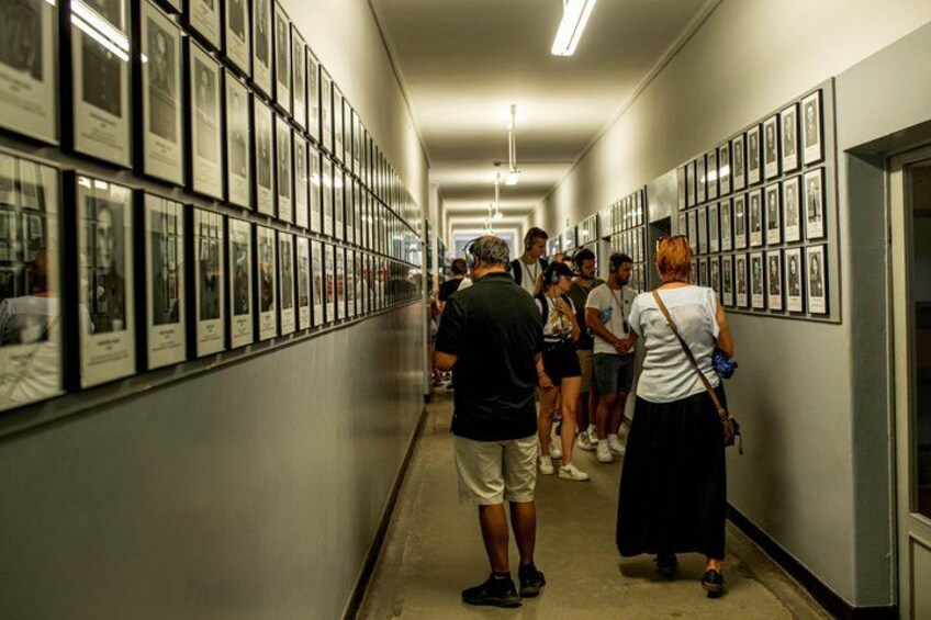 A hallway with framed pictures on the walls, people walking and standing, and a door at the end. 2