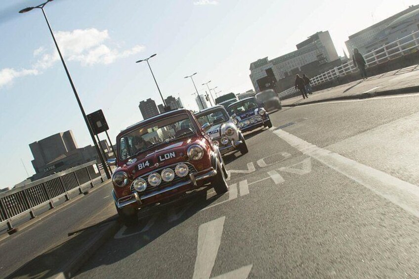 A red vintage car with 'BI4 LON' on the front, driving on a city street with modern buildings in the background. 3