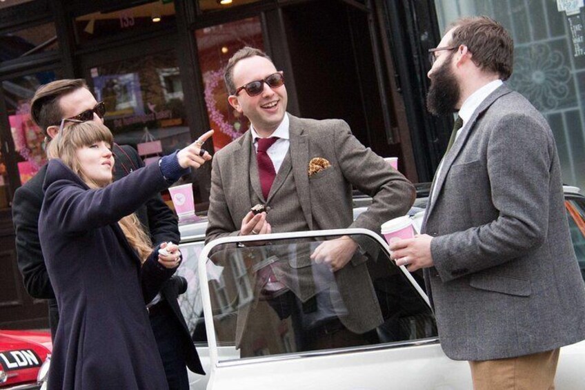 A man in a suit standing next to a car, holding a phone and a cup. 3