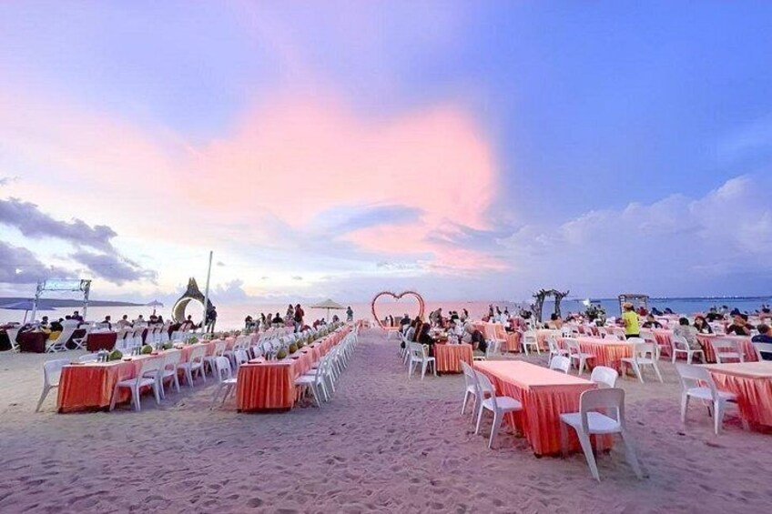 A beach wedding setup with rows of tables and chairs, a heart-shaped decoration, and a sunset in the background. 2