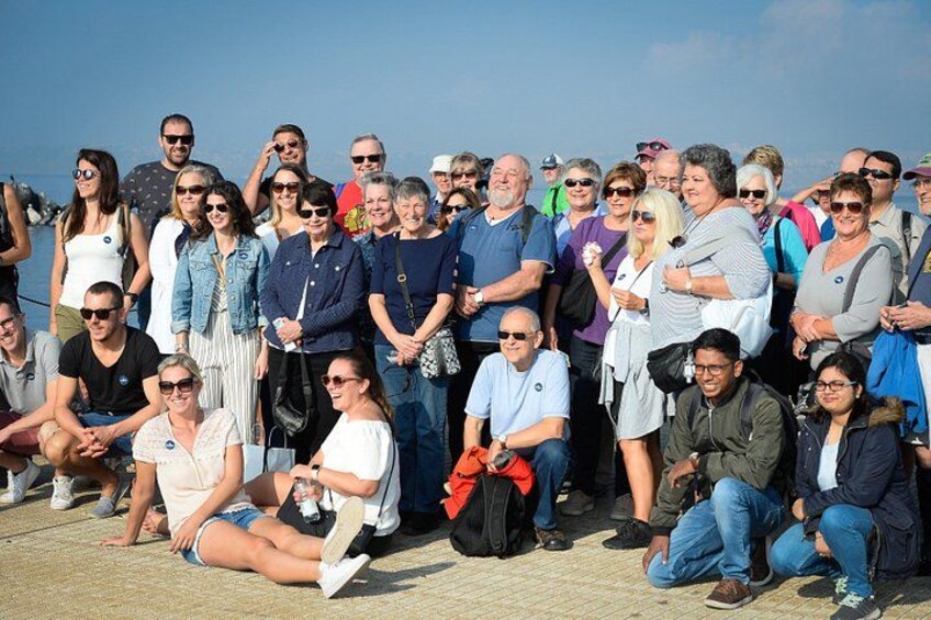 A group of people sitting on a concrete surface outdoors, with a body of water and a clear sky in the background. 3