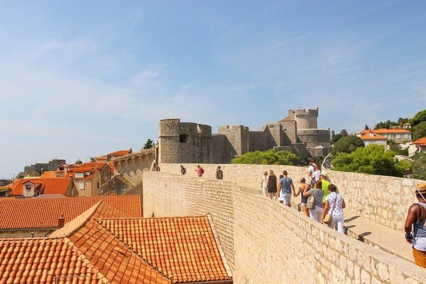 Un muro de piedra con gente caminando a lo largo, un castillo al fondo y techos de tejas rojas. 2