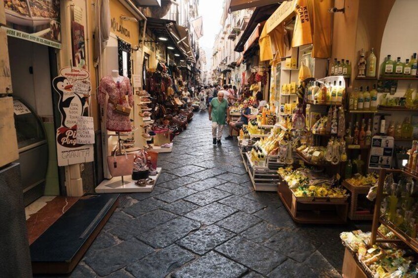 A narrow street with shops on both sides, including a shoe store and a market stall with fruits and vegetables. 2