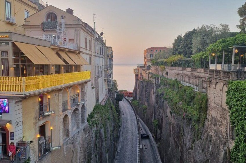 A coastal road with buildings on either side, a cliffside, and a sunset in the background. 4