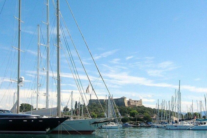 A marina with multiple sailboats docked, a clear sky, and a building on a hill in the background. 5