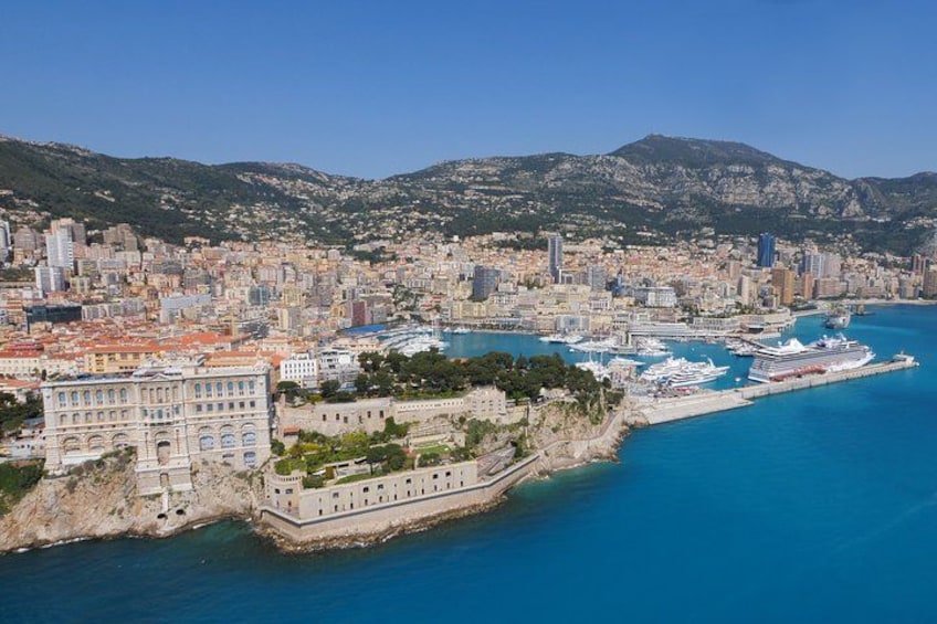Paesaggio costiero con un edificio storico imponente, un porto turistico con yacht e montagne sullo sfondo. 3