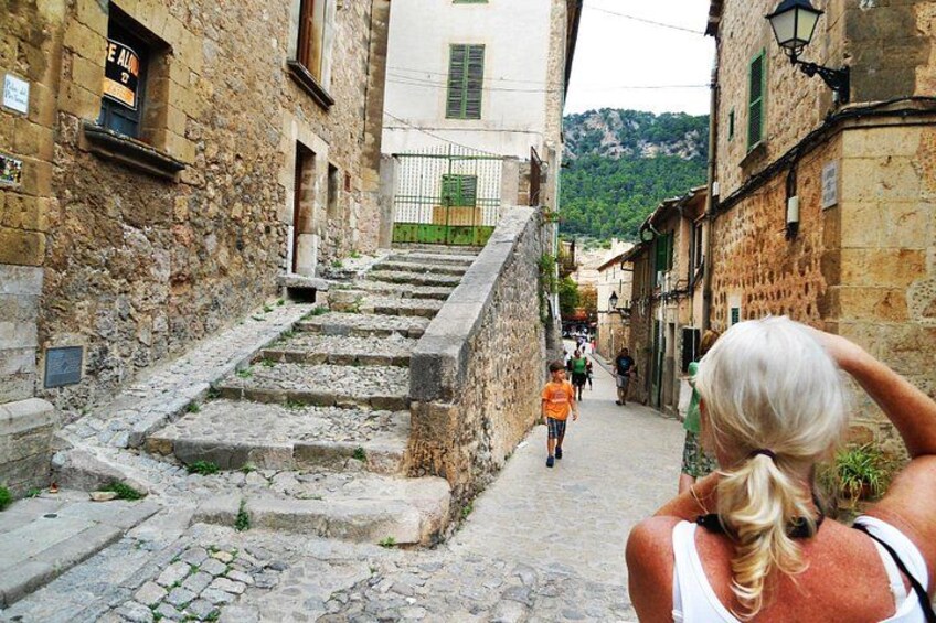 A narrow stone street with steps leading up to a building, surrounded by old stone buildings and greenery. 2