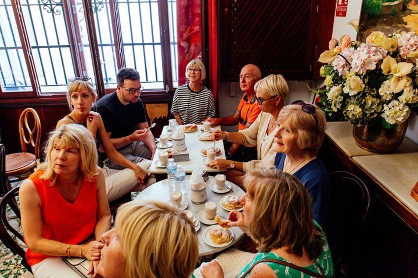 A group of people sitting around a table with food and drinks, in a room with windows and a vase of flowers. 3
