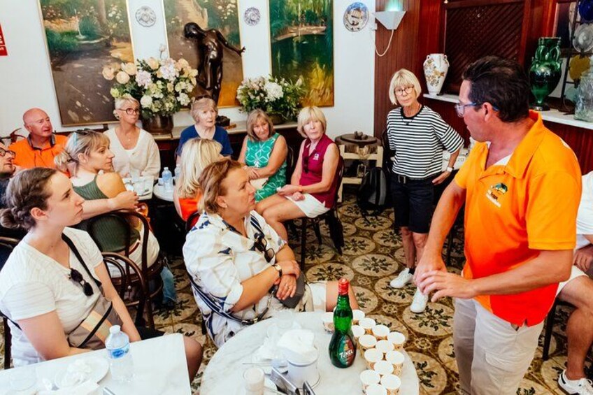 A group of people seated at tables in a room with floral decorations and artwork on the walls. 4