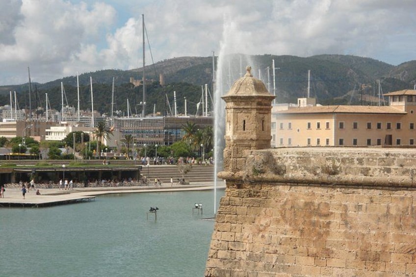 Une promenade en bord de mer, avec un mur de pierre d’époque, une fontaine et un bâtiment moderne, un mât sur lequel flotte un drapeau visible en arrière-plan. 3