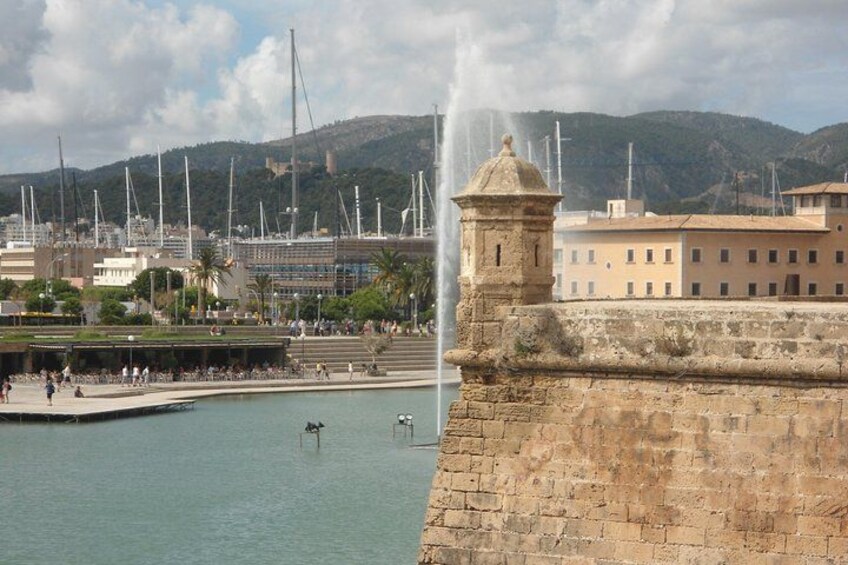 A coastal cityscape with a historic stone wall, a fountain, and a marina with sailboats. 5