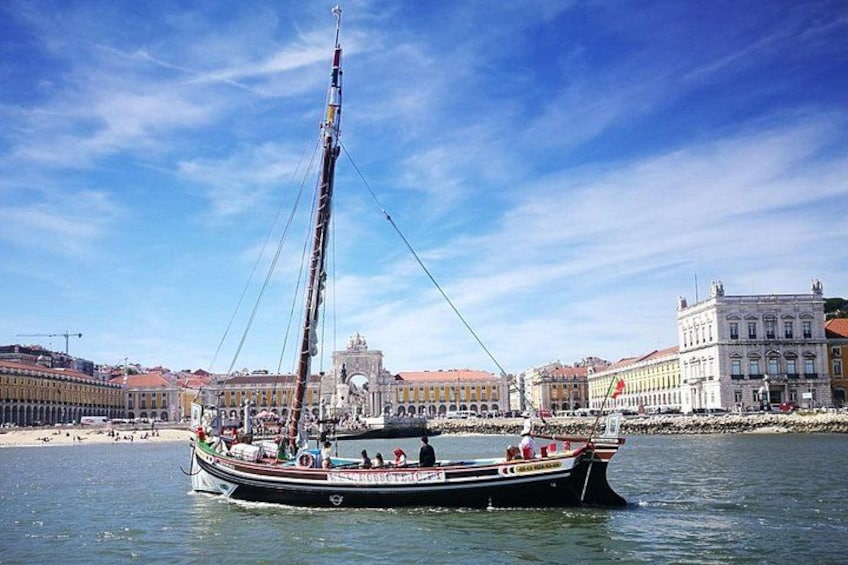 A traditional sailing boat with a tall mast, anchored near a waterfront with buildings in the background. 1