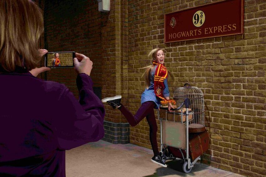 Eine Person fotografiert eine andere Person, die einen blauen Mantel und einen roten Schal trägt und neben einem Käfig mit einem Vogel steht. An der Wand hängt ein Schild mit der Aufschrift 'Hogwarts Express'. 5