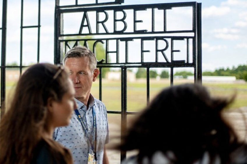 A man and a woman standing in front of a gate with the words 'ARBEIT MACHTEREI' visible. 3