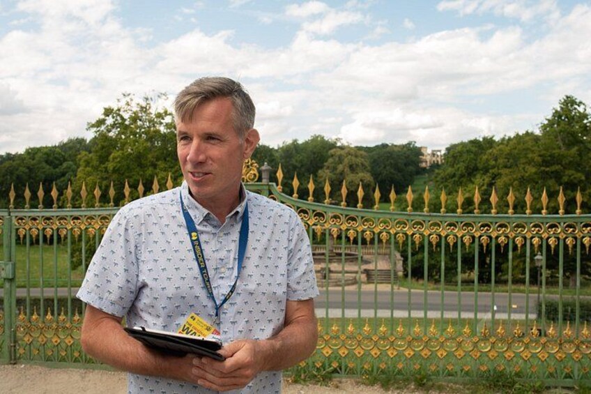 A man holding a clipboard standing in front of a decorative fence with a park in the background. 5