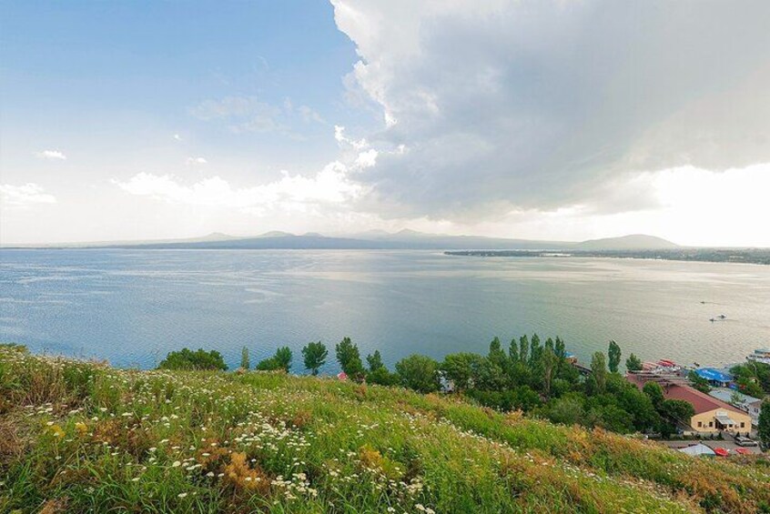 Un lago con una cadena montañosa distante, un primer plano cubierto de hierba y flores silvestres, y algunos edificios a la derecha. 5