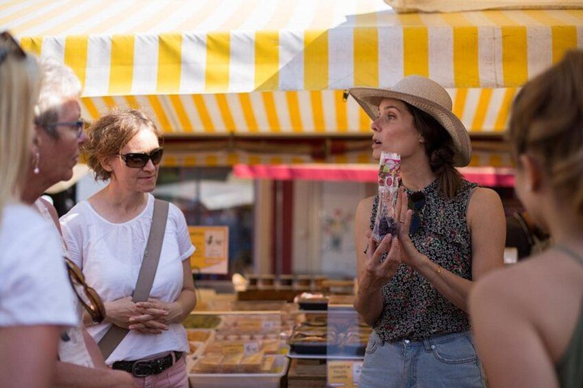 Un puesto de mercado con toldo a rayas, gente mirando productos y una mujer sosteniendo un artículo. 5
