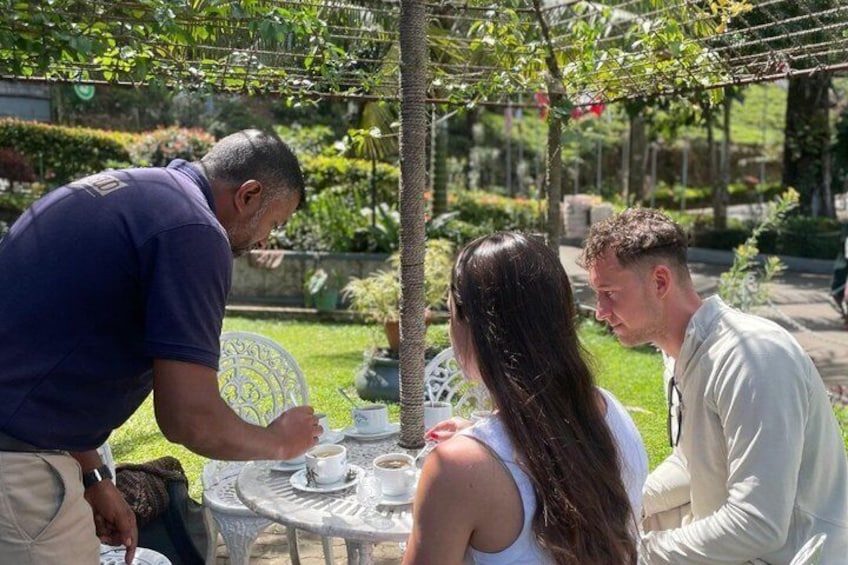 A man serving coffee to a woman and a man at an outdoor table with a garden in the background. 1