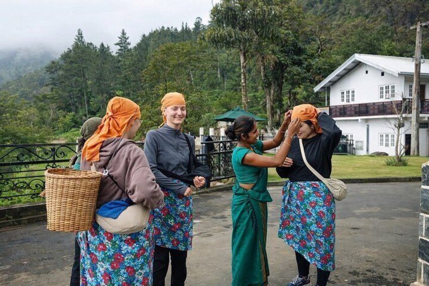 Fyra personer klädda i traditionella dräkter, varav en hjälper en annan att fästa en huvudduk. Bilden är tagen på landsbygden med ett hus och grönska i bakgrunden. 5