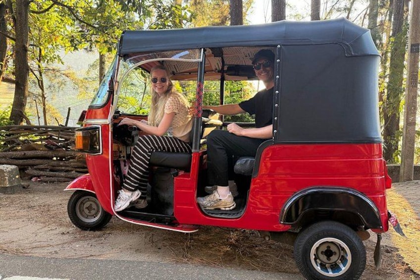En rød tuk-tuk med to passagerer, hvoraf den ene kører og den anden sidder, på en støvet sti omgivet af træer. 4