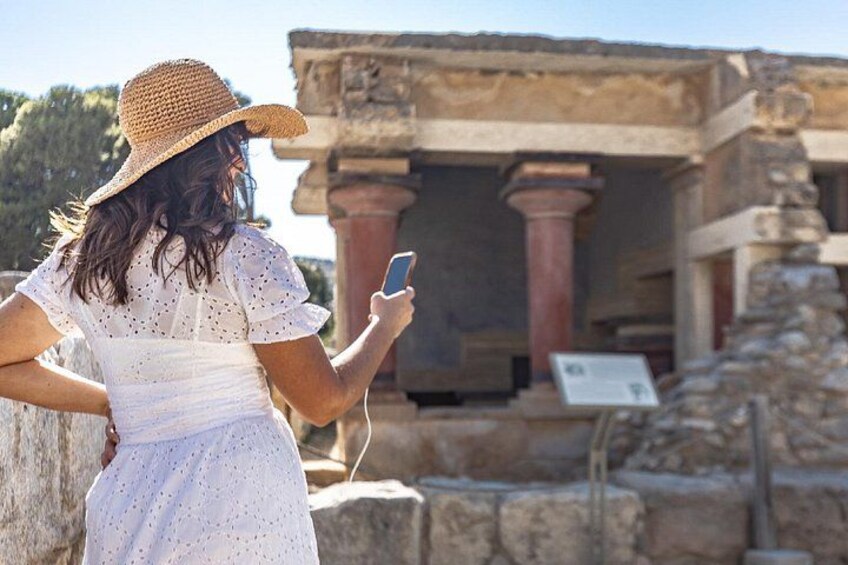A person in a white dress and straw hat using a smartphone, standing in front of ancient ruins with columns and stone blocks. 1
