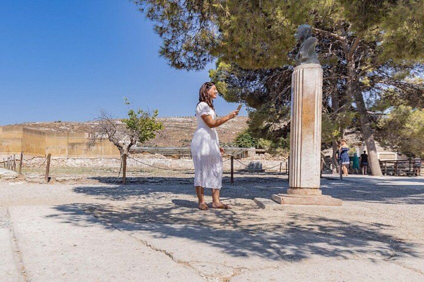A person in a white dress standing near a column with a statue on top, in a sunny outdoor area with trees and a clear sky. 3