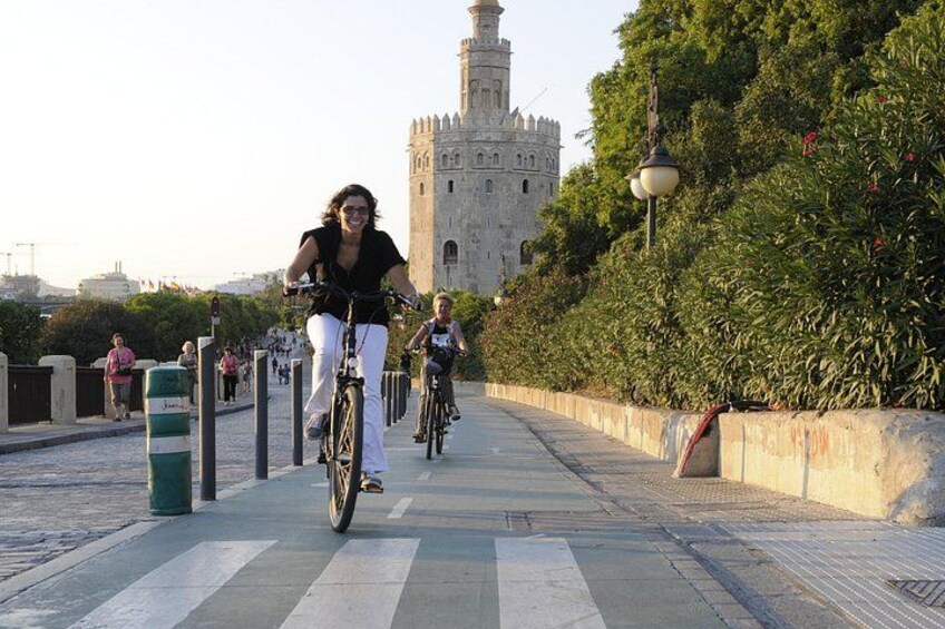 A paved pathway with cyclists, a historic tower, and a green hedge. 1