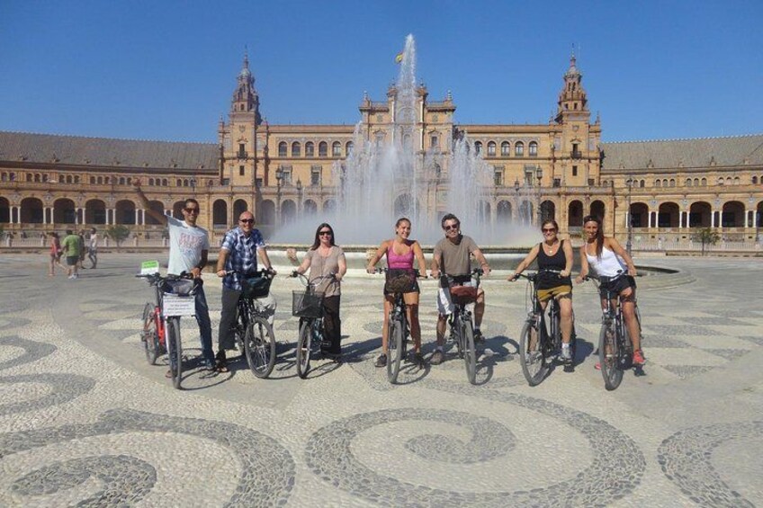 A group of people on bicycles in front of a historic building with a fountain. 1