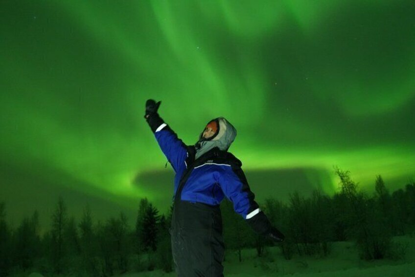 A person wearing a blue jacket and a helmet, standing in a forest with green aurora lights in the sky. 4