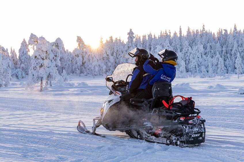 A snowmobile with two riders on a snowy landscape with trees in the background. 1