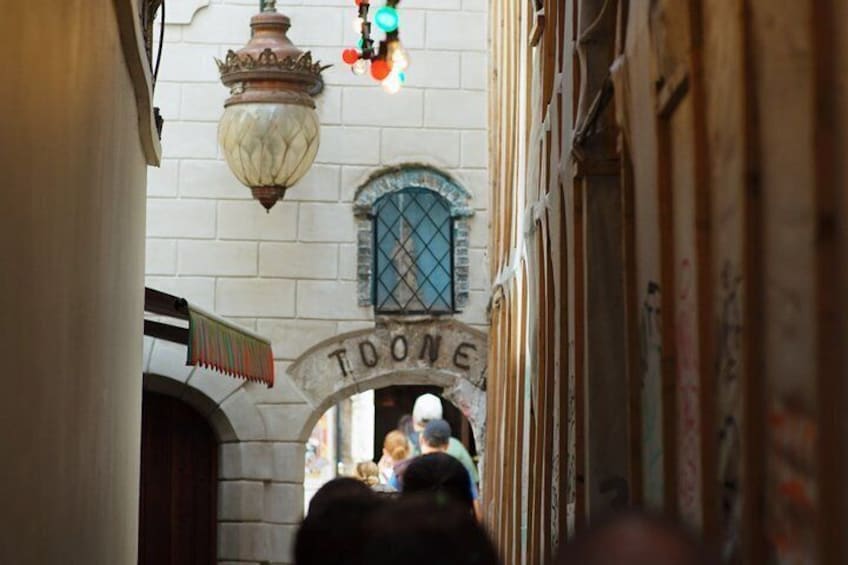 A narrow alleyway with a 'TOONE' sign above an archway, a hanging lantern, and a person walking away from the camera. 3