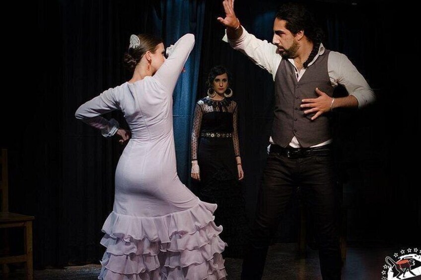 A person in a ruffled dress dancing, another person in a vest and shirt gesturing, and a third person in a patterned dress in the background. 2