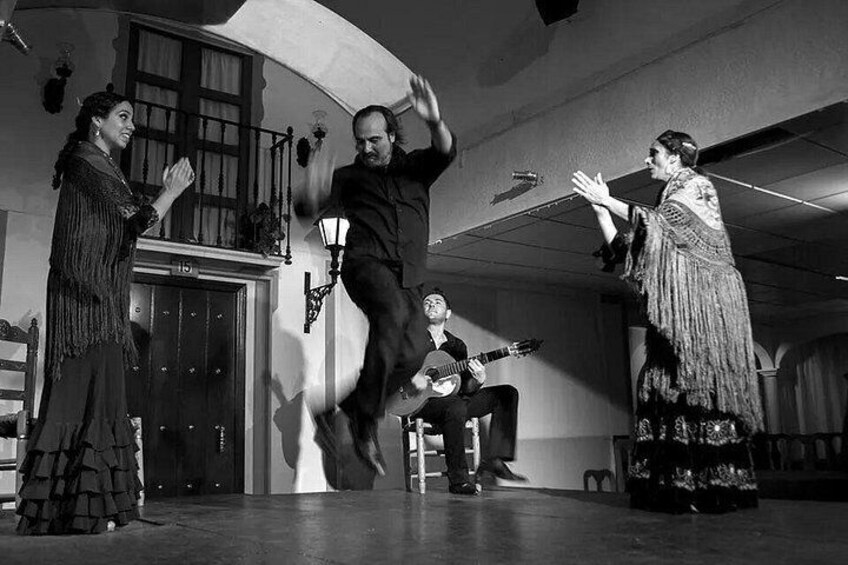 A black and white photo of a stage with three people, one playing guitar, another dancing, and a third clapping. 4