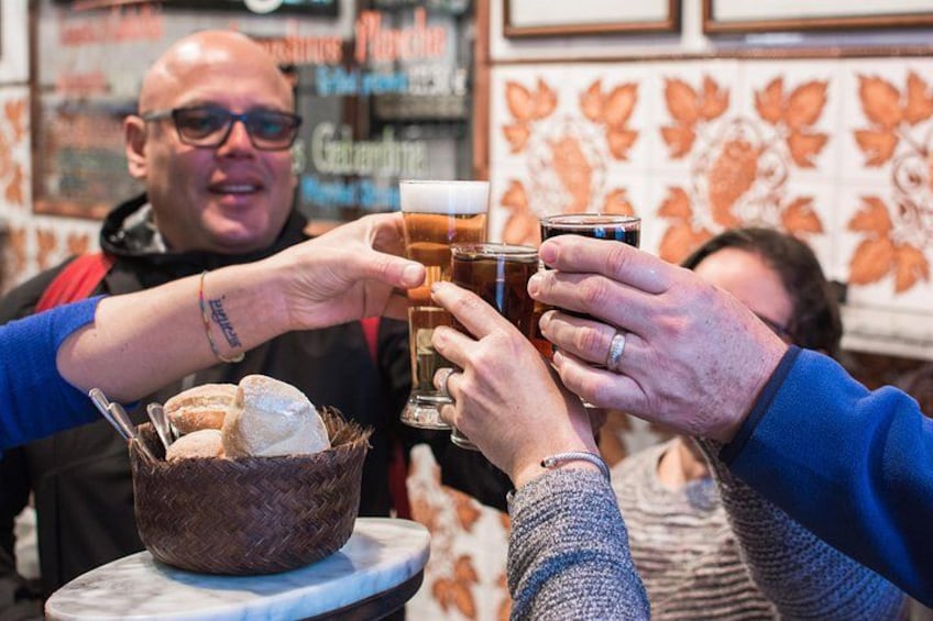 Personas brindando con vasos de cerveza, una cesta de pan y una pared de azulejos con diseños florales. 4