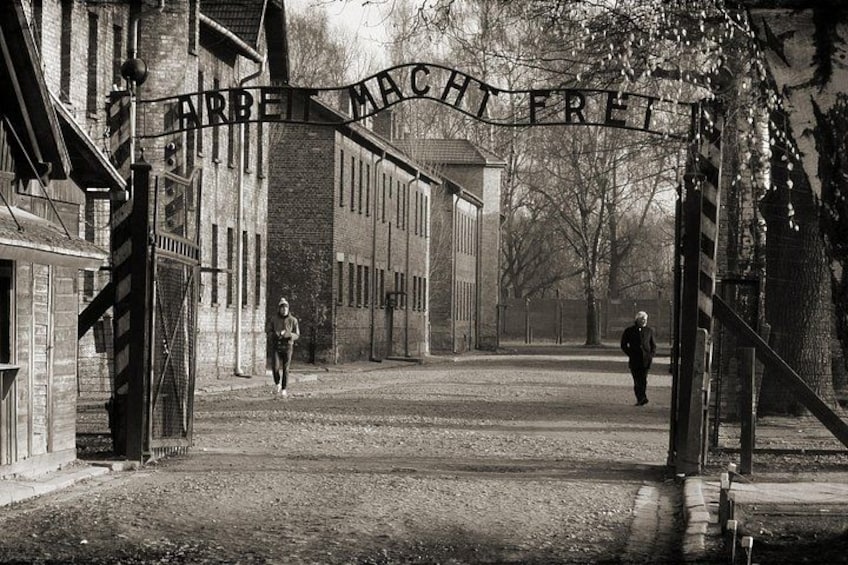 A historical building with a sign reading 'ARBEIT MACHT FREI' and two people walking in the foreground. 1