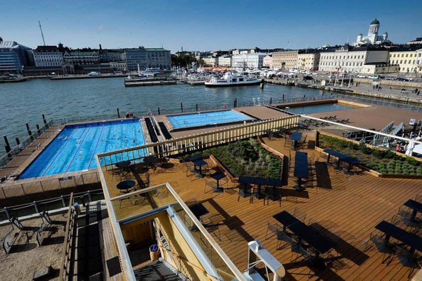 A cruise ship deck with a swimming pool, lounge chairs, and a view of the city and water. 2