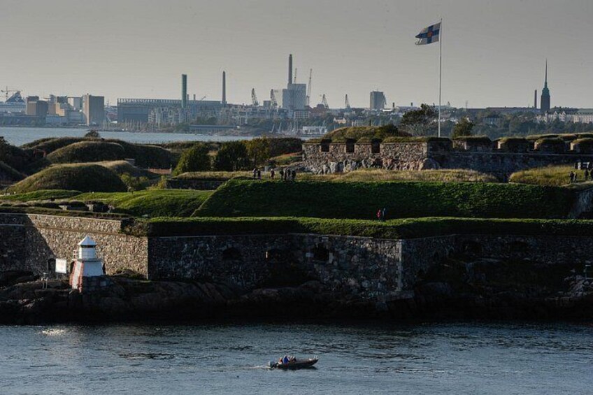 A coastal fortification with green grassy mounds, a lighthouse, and a city skyline in the background. 5