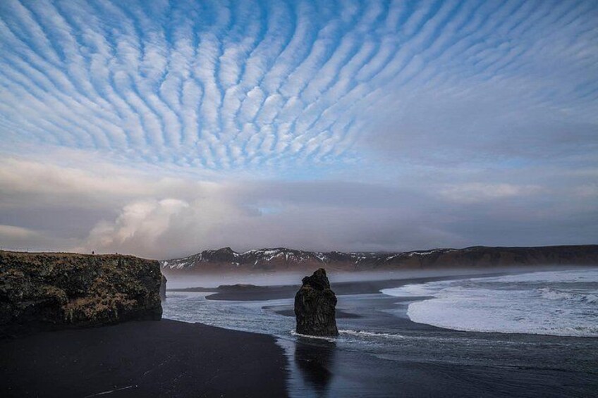 Un paisaje costero con una formación rocosa destacada, una playa de arena y montañas nevadas en la distancia bajo un cielo con nubes. 5