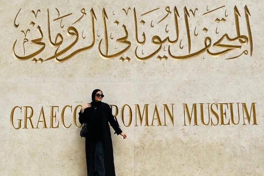 Een persoon staat voor het bord van het Graeco-Romeins Museum, met Arabische tekst erboven. 1