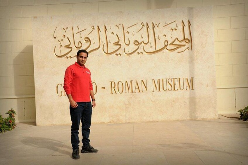 A person standing in front of a wall with Arabic and English text, including 'Roman Museum'. 5
