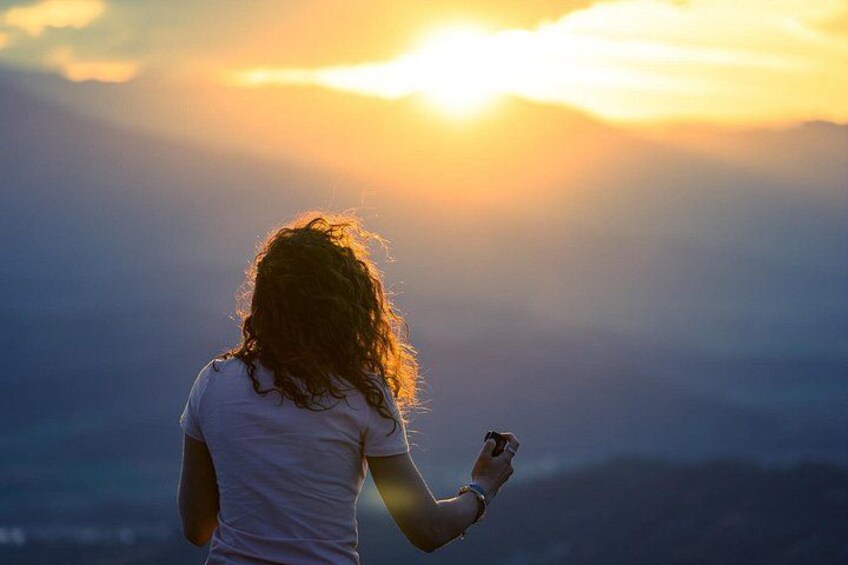 Une personne aux cheveux bouclés, vêtue d’une chemise blanche, se tient de dos à la caméra et surplombe un paysage montagneux au coucher du soleil. 4