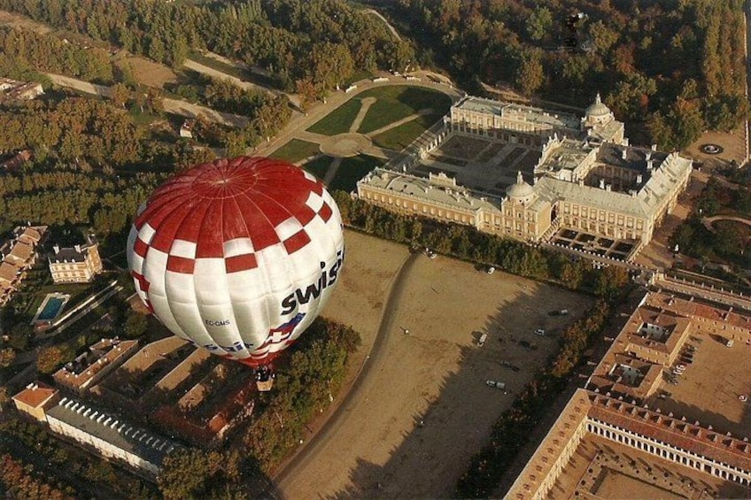 A hot air balloon with 'SWISS' written on it flying over a large, historic building surrounded by trees and open spaces. 3
