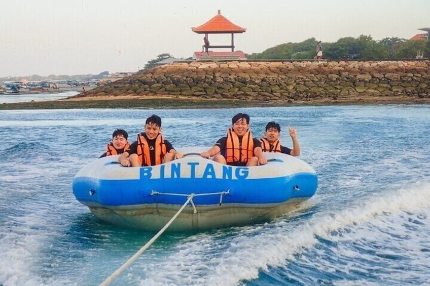 A group of people in a blue and white inflatable boat labeled "BINTANG" on a body of water with a rocky shore and a gazebo in the background. 5