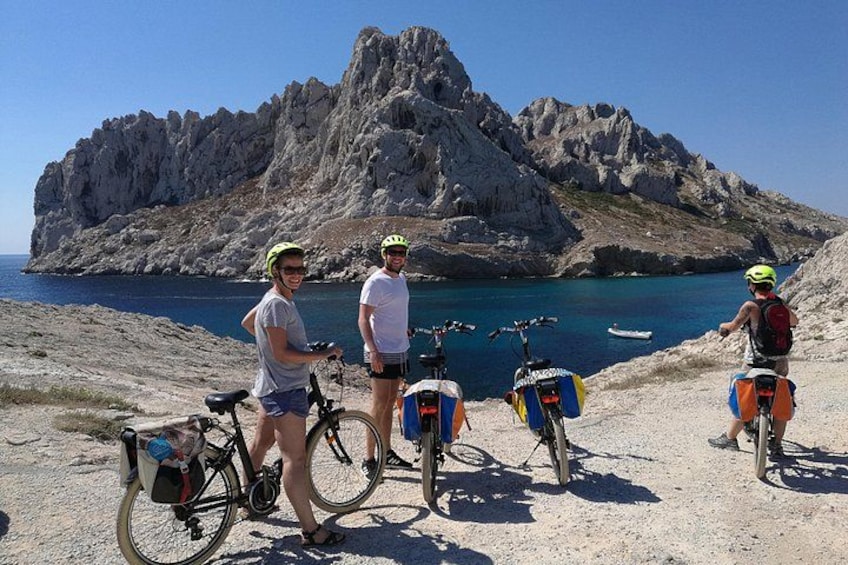 Three cyclists with bicycles on a coastal path, rocky cliffs, and a clear blue sea in the background. 1