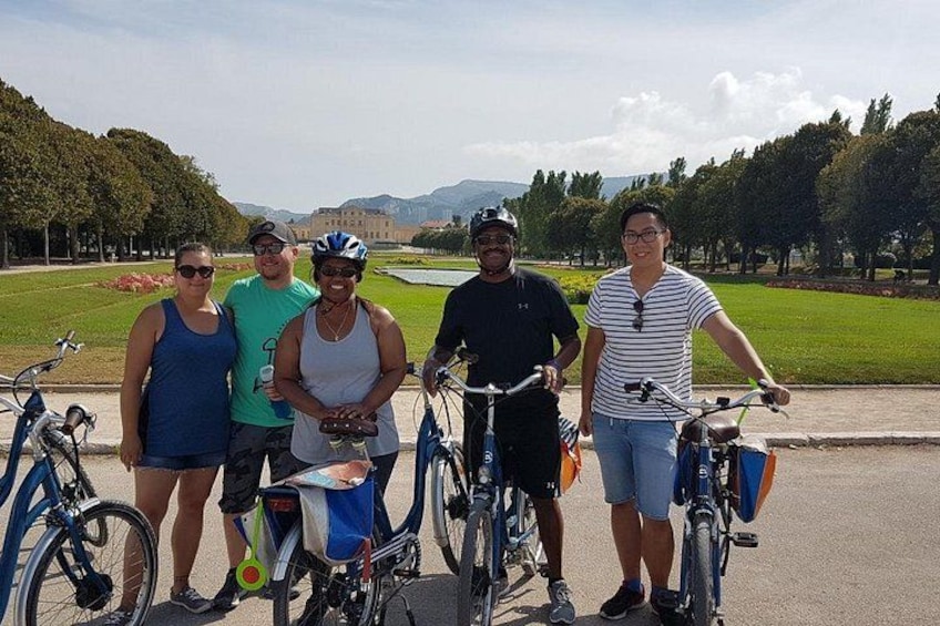 A group of people with bicycles in a park with trees and a building in the background. 2