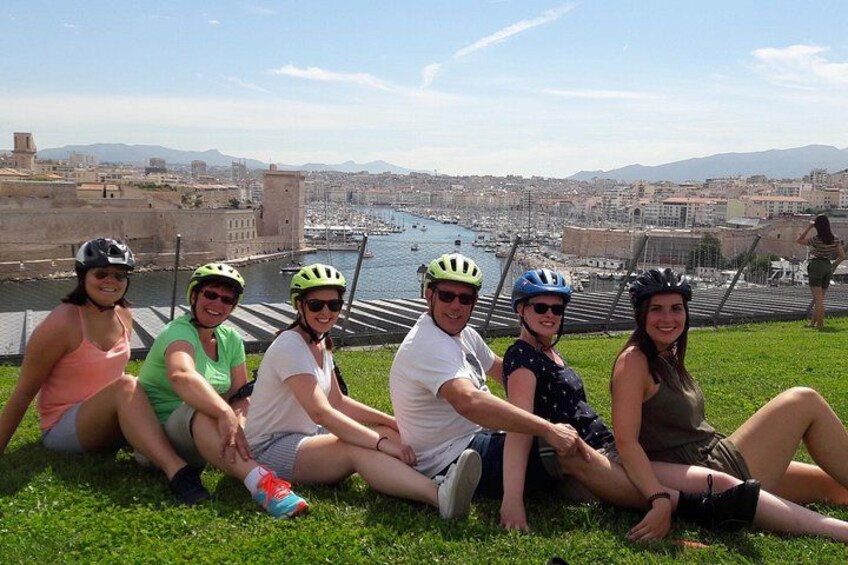 A group of people sitting on grass with a cityscape and water in the background. 3