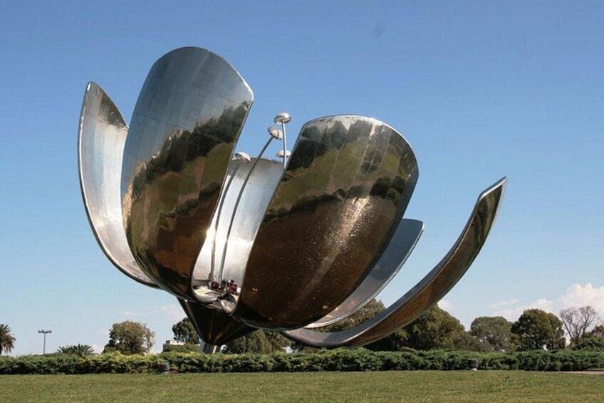 Een groot metalen sculptuur, vormgegeven als een bloem met bloemblaadjes, in een grasveld met bomen op de achtergrond. 3