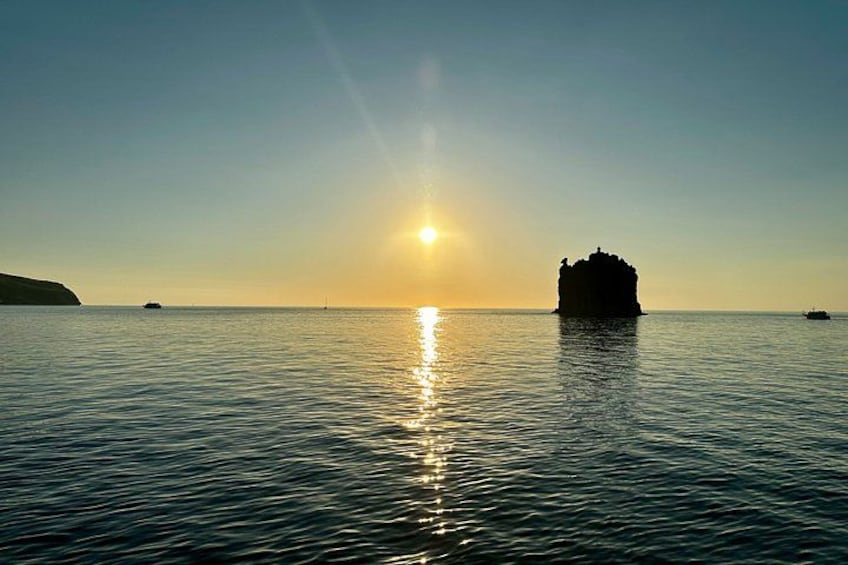Une mer calme au coucher du soleil, avec une formation rocheuse imposante au centre et une masse terrestre lointaine sur la gauche. 3